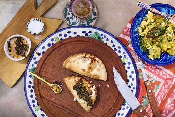 Image of a wooden plate with two empanadas on it, there are several other items on the photo: fork/knife, bowl of potato salad and a beverage. 